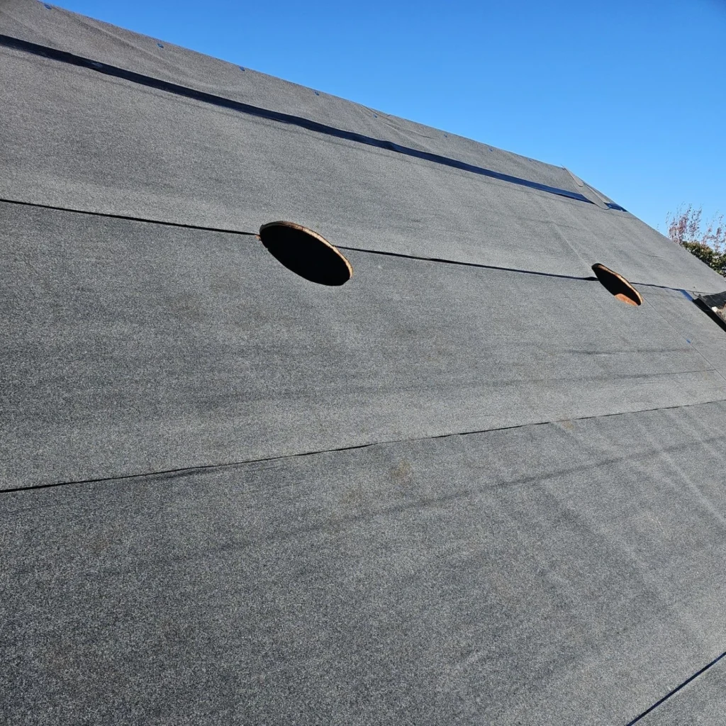 Gray roofing underlayment installed on residential roof with two circular pipe boot cutouts awaiting vent installation, blue sky background.
