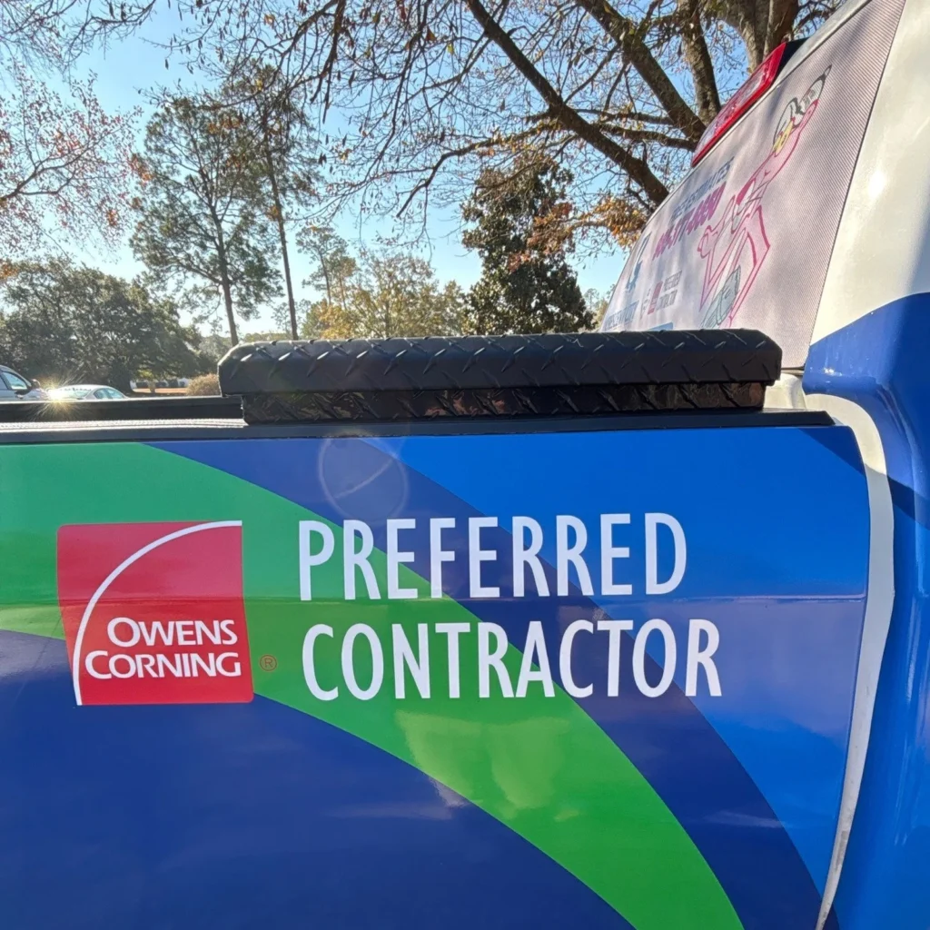 Owens Corning Preferred Contractor logo on blue and green branded truck tailgate, parked outdoors under trees.