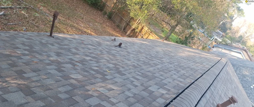 Gray architectural shingle roof replacement with pipe vents, ridge cap, and gutters on residential home near wooded hillside, Slidell