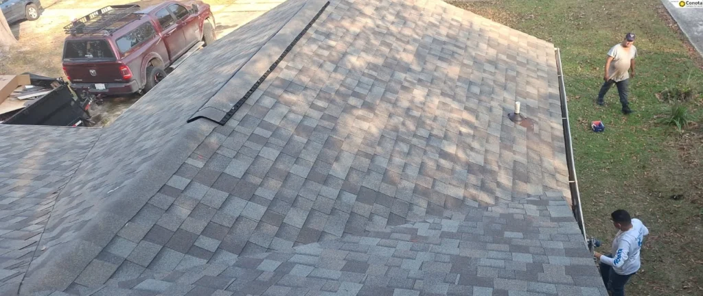 Aerial view of completed architectural shingle roof replacement with gutters and pipe boot vent, two workers on ground below.
