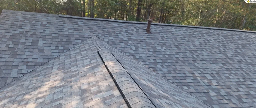 Gray architectural shingles on residential roof with hip ridge, pipe vent, and wooded background in Slidell
