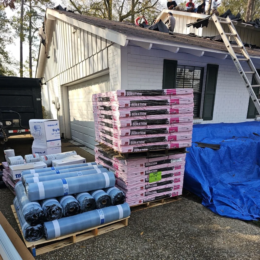 Pallets of Owens Corning Duration shingles and roofing underlayment rolls staged in driveway as crew tears off old roof on large Mandeville LA home.