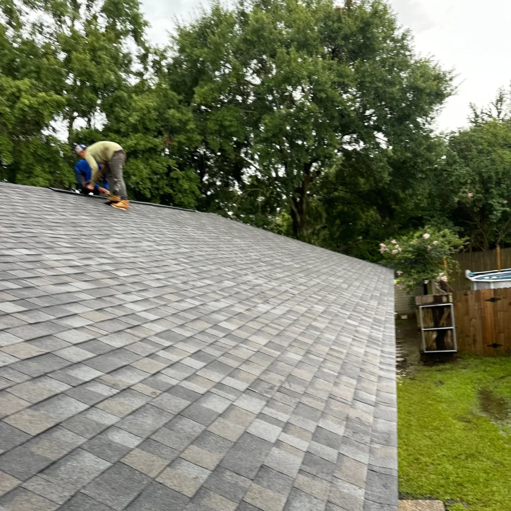 Roofers installing OC Williamsburg Gray asphalt shingles on large residential roof slope in Slidell, lush trees in background.